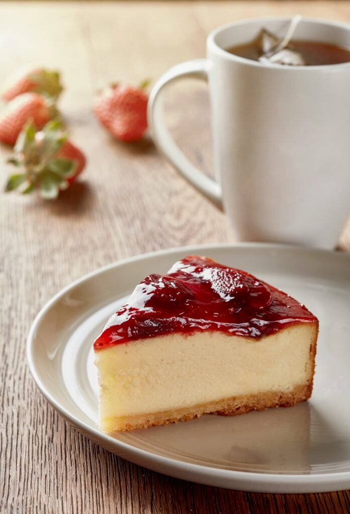 Porción de cheesecake de fresas sobre plato beige con taza de té detrás y fresas de fondo